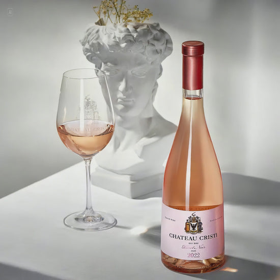 Sticla Chateau Cristi Pinot Noir Rose 2022 langa un pahar de vin rose, pe fundal bust clasic alb